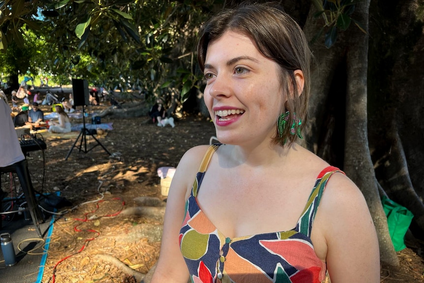 A young woman wearing bright green earrings and patterned dress stands under a fig tree