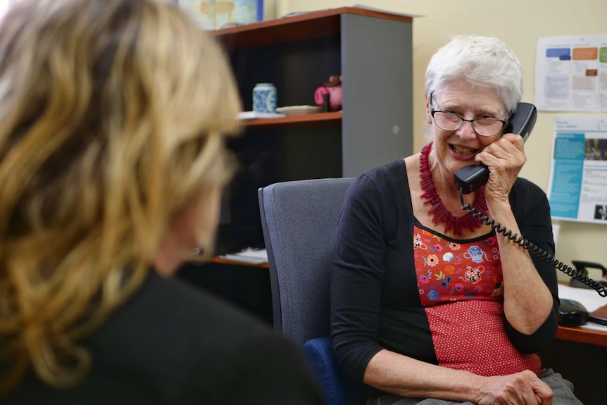 An elderly woman with short, grey hair speaks on a telephone.