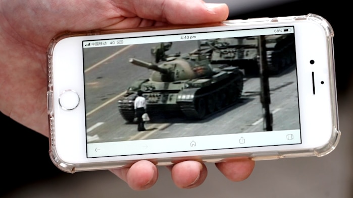 A hand holding an iPhone with the image of a man standing in front of tanks on the screen.