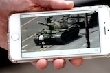 A hand holding an iPhone with the image of a man standing in front of tanks on the screen.