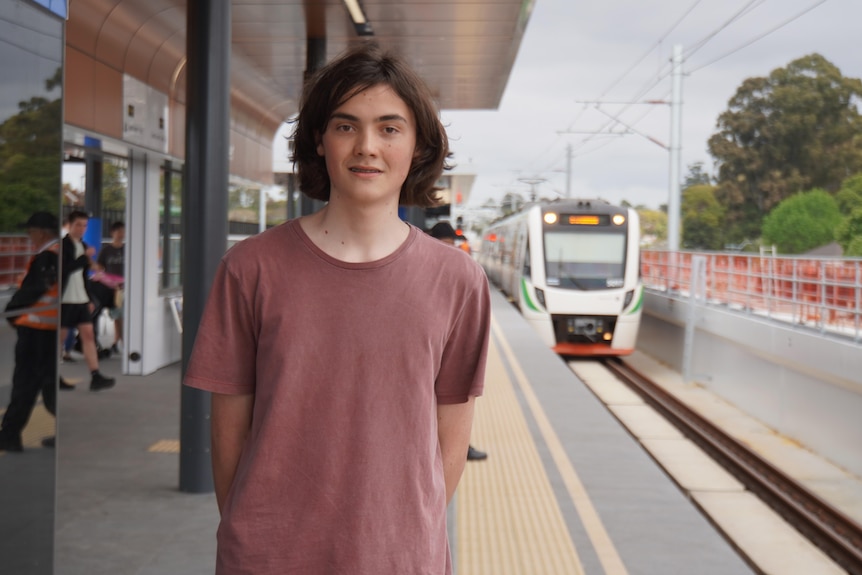 Un adolescente de pelo largo y oscuro sonríe a la cámara en una estación de tren