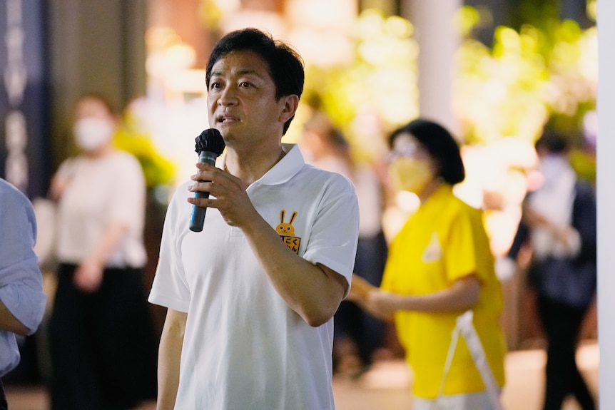 A man with dark hair and wearing a white shirt holds a microphone as he stands in the middle of a room.