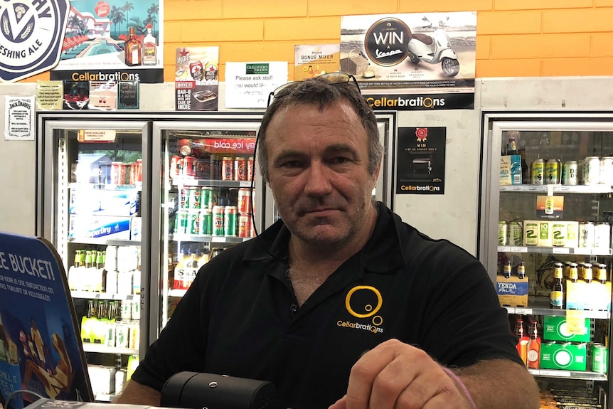 A middle-aged man in a black shirt stands behind the counter at a bottleshop