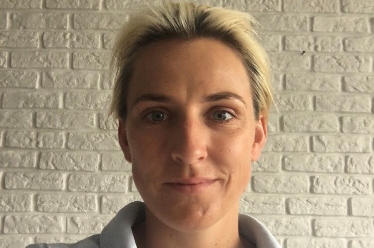 Selfie of a woman with short blonde hair wearing a white polo shirt in front of a cream brick wall.