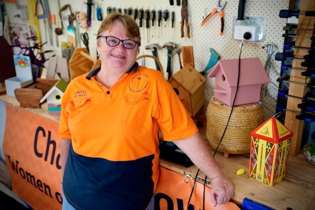 Woman standing in front of tool bench, wearing an orange high vis shirt.