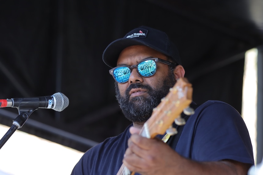 A Torres Strait Islander man with sunglasses and hat performs on stage with guitar