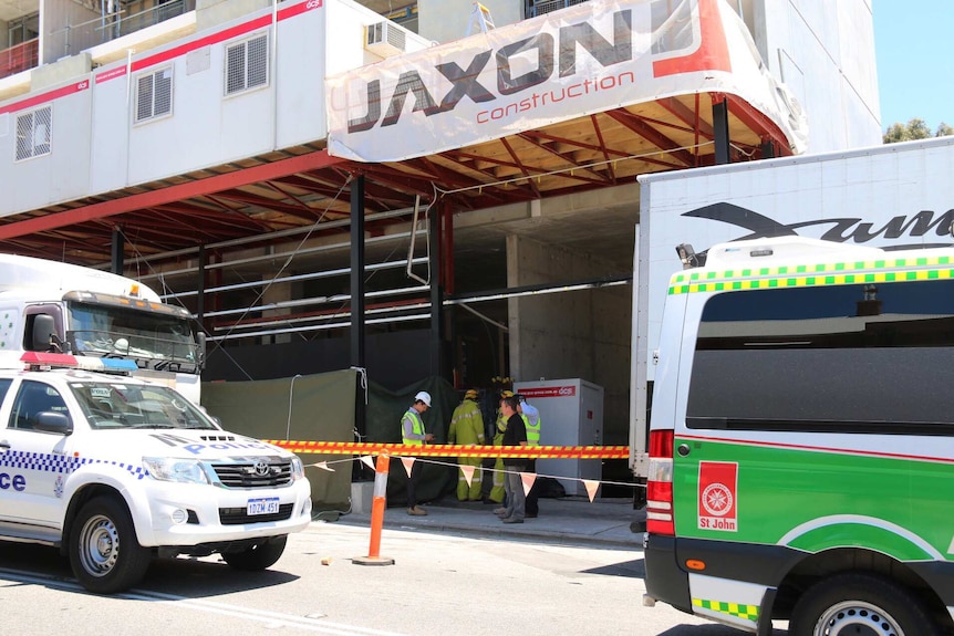 A police car and an ambulance sit parked outside a building site as workers stand at the entrance.