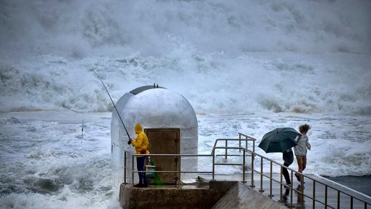 A fisherman in yellow wet weather gear takes shelter behind a small building beside the ocean as large waves crash around him.