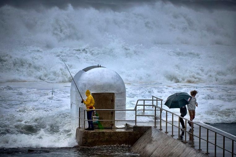 A fisherman in yellow wet weather gear takes shelter behind a small building beside the ocean as large waves crash around him.