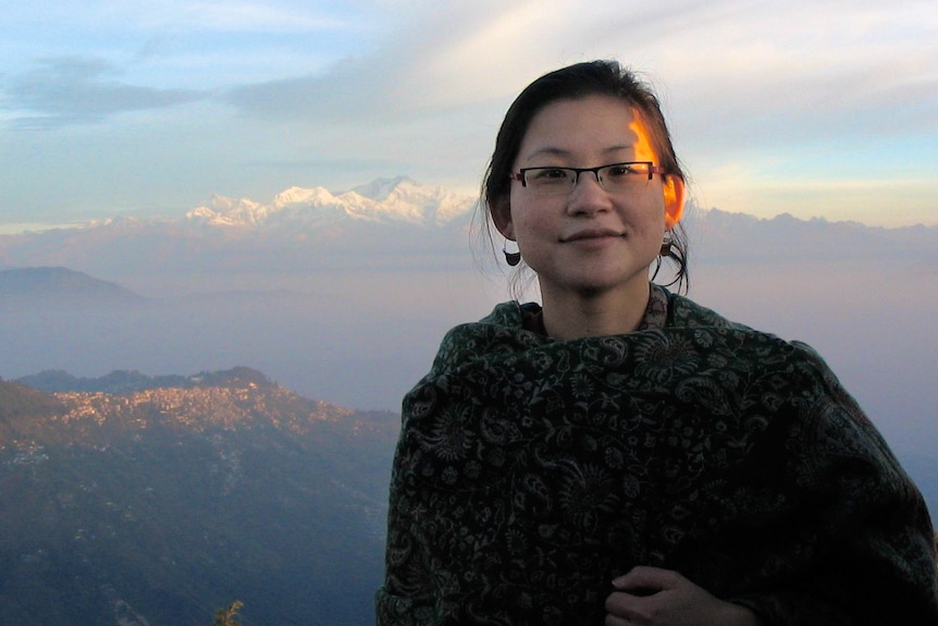 A woman in a green shawl stands in front of a wide mountain range at sunset. 
