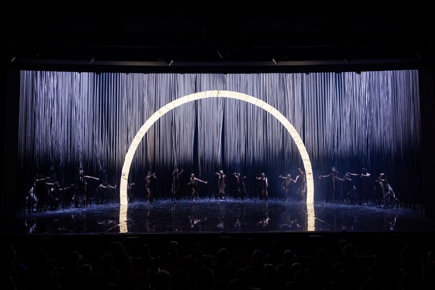 A darkened stage with a line of dancers posing behind an illuminated arch positioned centre-stage.