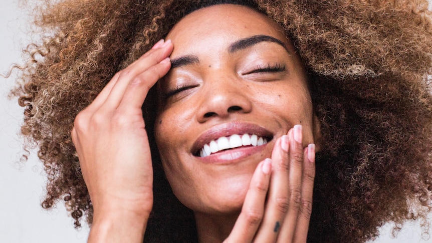 A woman rubs moisturiser on her face, smiling.