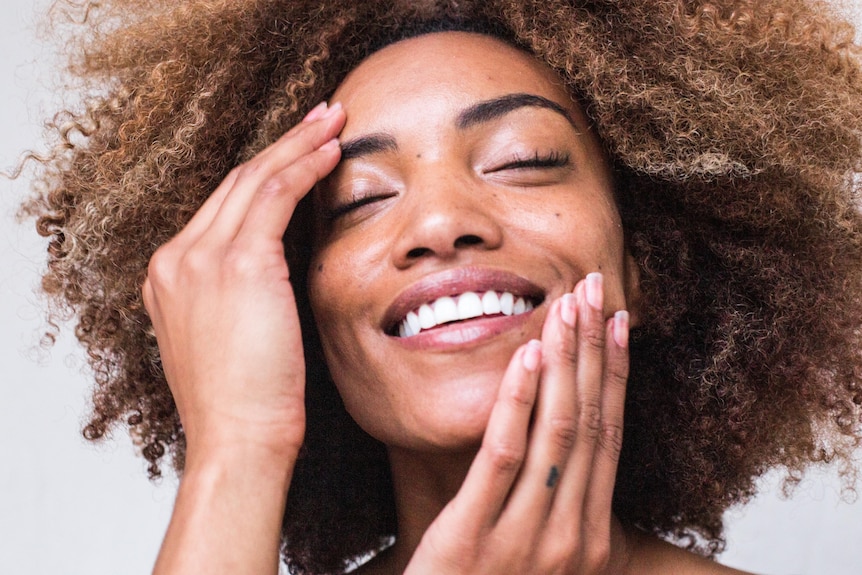 A dark skinned woman puts moisturiser on her face, smiling