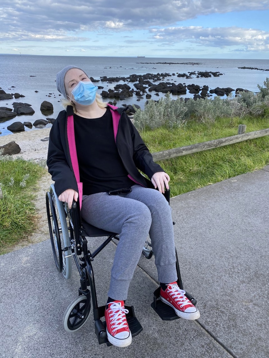 A woman in a wheelchair wearing a beanie and tracksuit poses for a photo by the sea