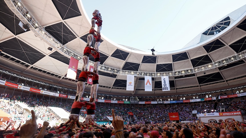 A group of people in red shirts form a large human tower in the middle of a round arena.