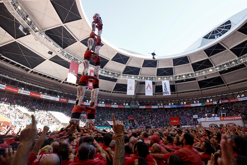 A group of people in red shirts form a large human tower in the middle of a round arena.
