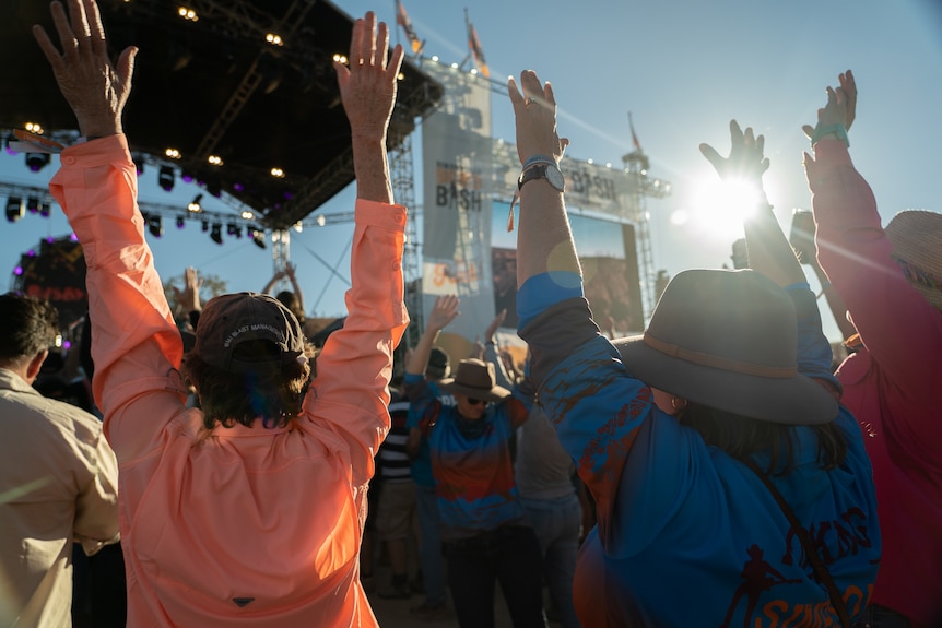 People with their arms up dancing in front of a stage. The photo is shot from behind them.