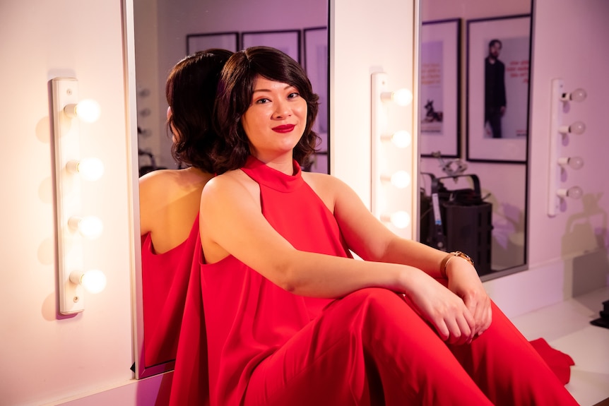 A 30-something Asian Australian woman sits on a dressing room table, her back against a mirror, at Belvoir St Theatre