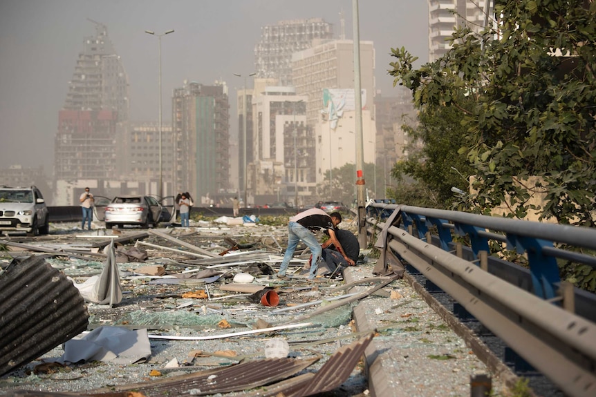 Two men sit amongst broken glass and strewn wreckage on a road with damaged buildings in the background.