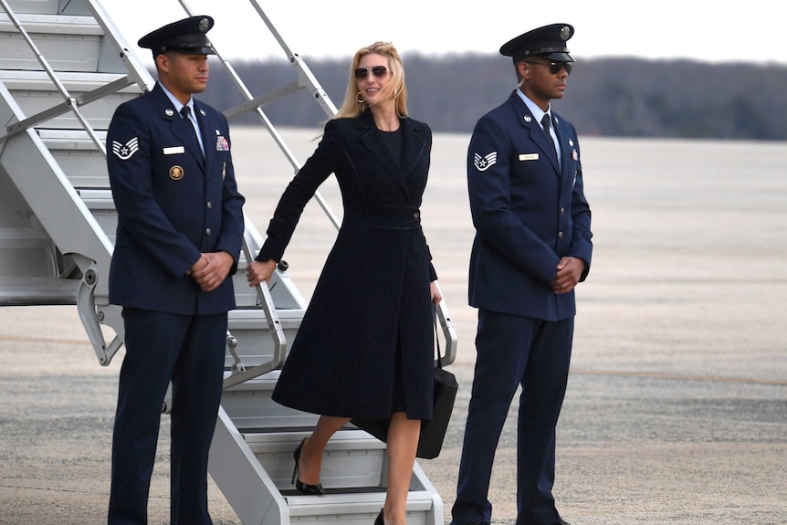 Ivanka Trump walks own a plane staircase, flanked by two men in military uniforms. She is wearing a dark blazer and sunglasses.