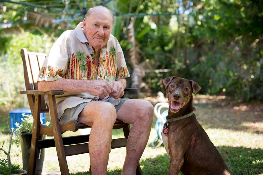 An older man sits in a garden chair with a large, red-brown dog sitting beside him.
