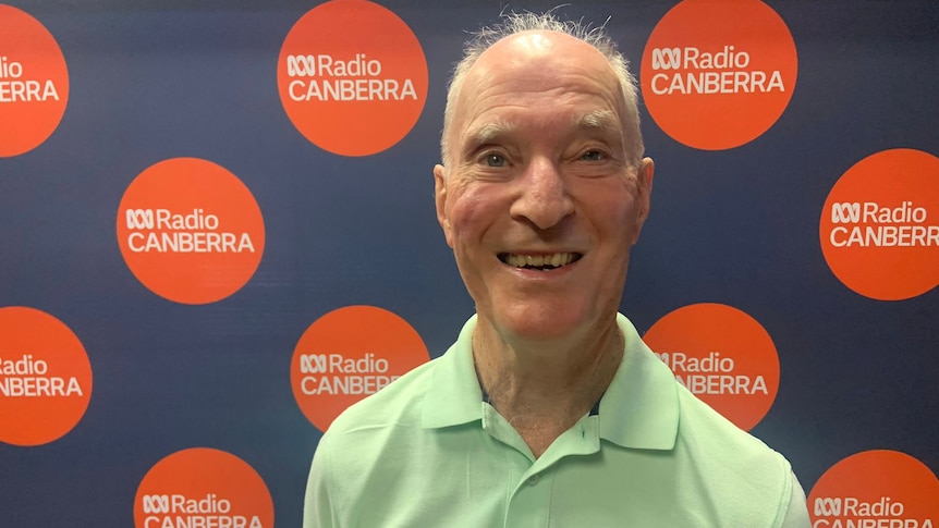 Graham stands smiling in a green shirt in front of the ABC Canberra logo