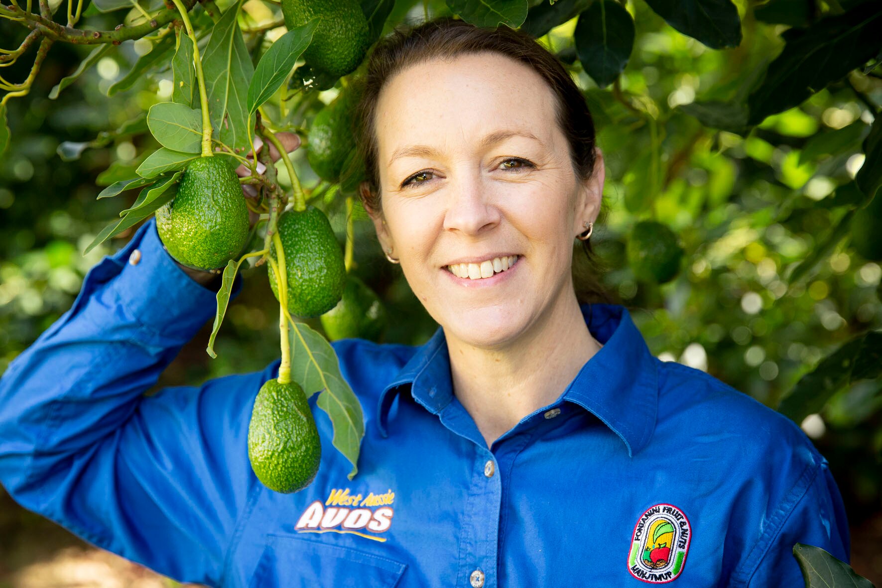 Una mujer con sonrisas de cabello castaño que se inclina en un árbol sosteniendo una rama con fruta verde con forma de ovalada colgando de ella.