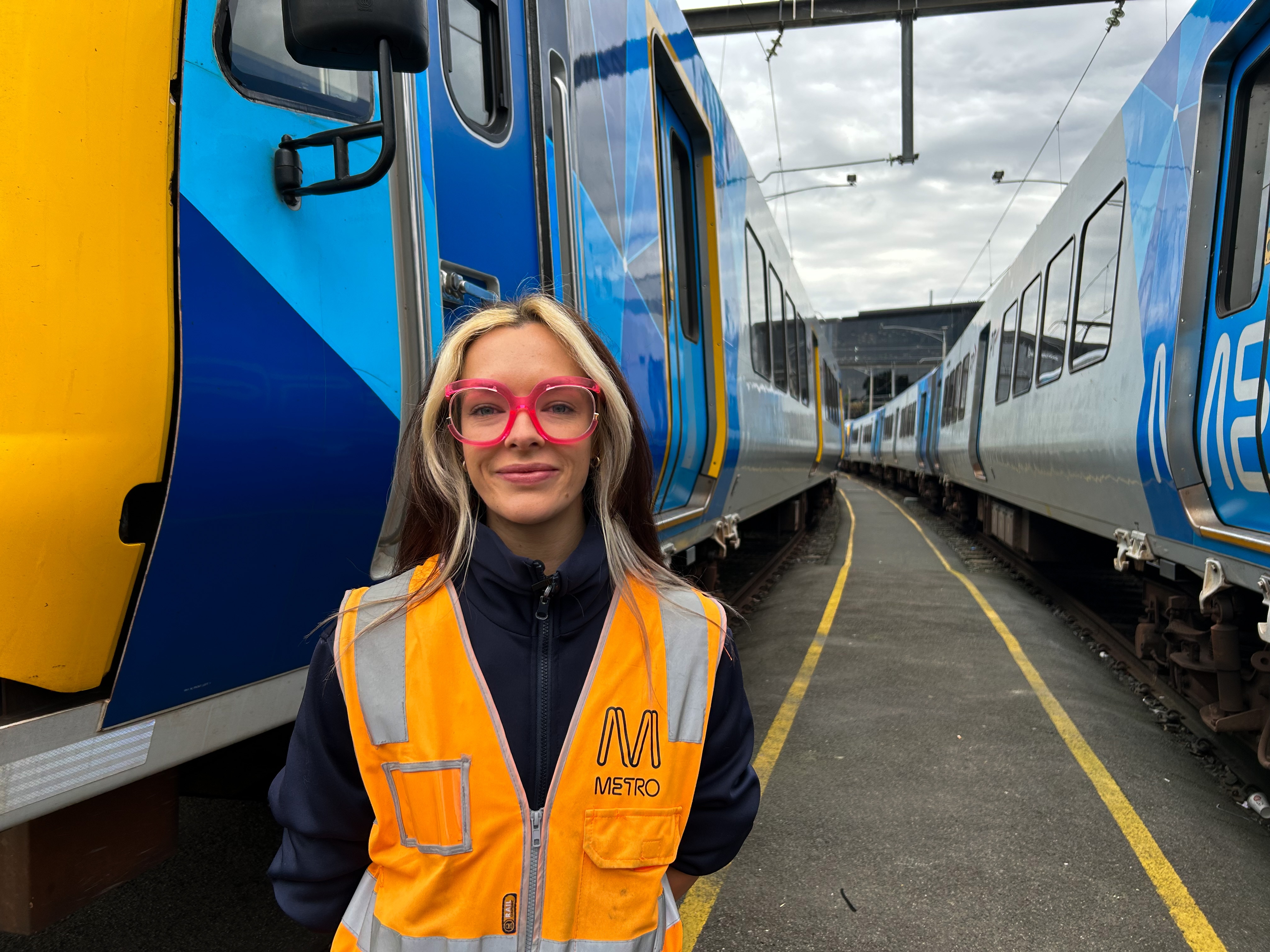 A young woman in a fluoro vest with pink glasses standing between trains.
