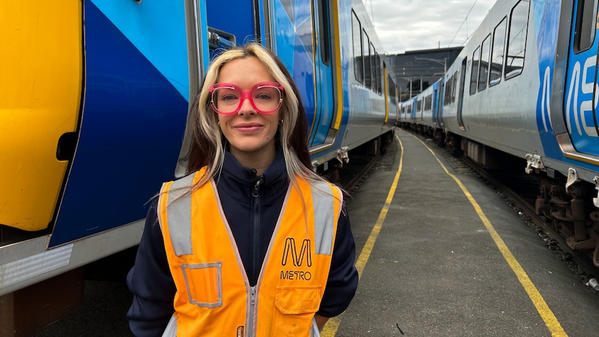 A young woman in a fluoro vest with pink glasses standing between trains.