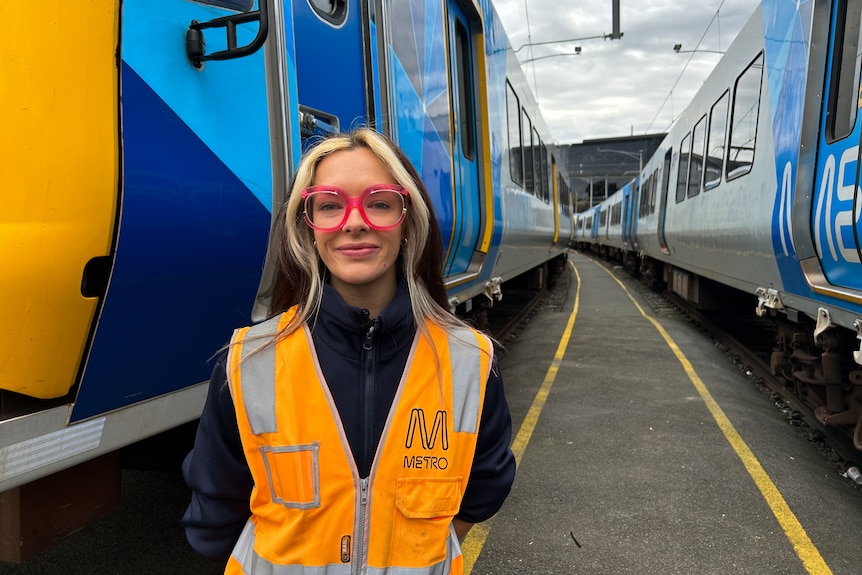 A young woman in a fluoro vest with pink glasses standing between trains.