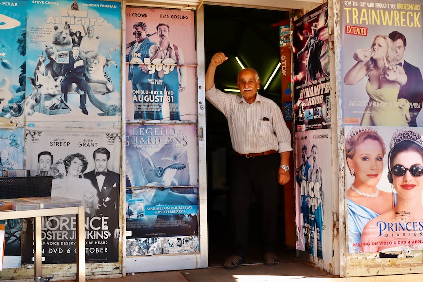 A man stands in the doorway of his video store with movie posters in the windows.