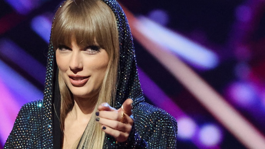 Blond woman in sparkly top points at camera against purple backdrop