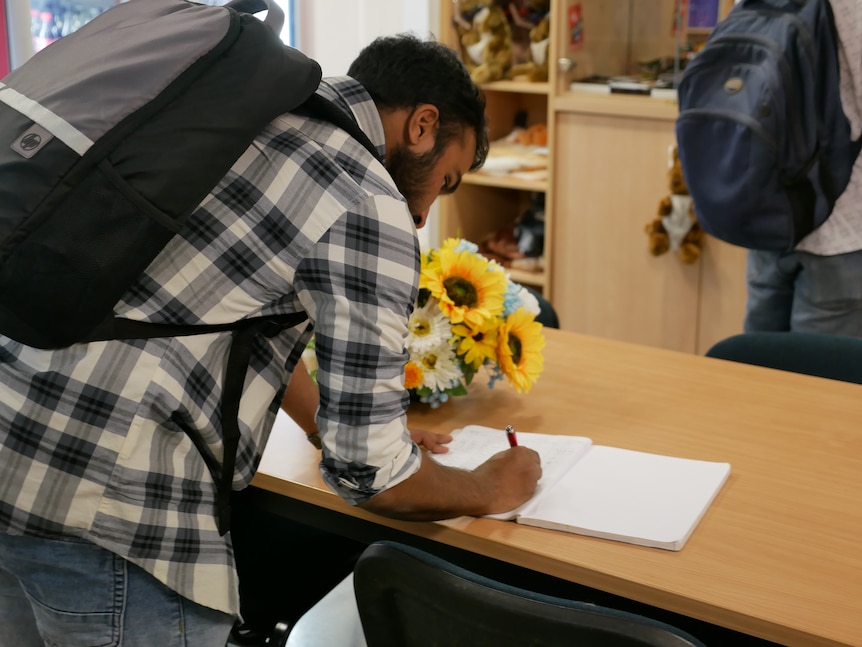 A man leans over a table to sign a paper booklet, next to a vase of flowers.