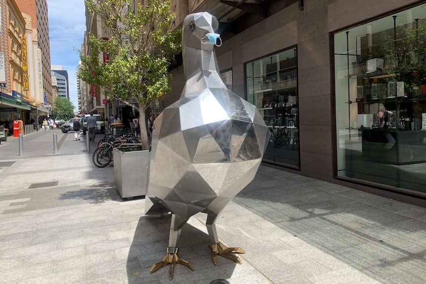 A silver statue of a pigeon wearing a blue surgical mask in a shopping area