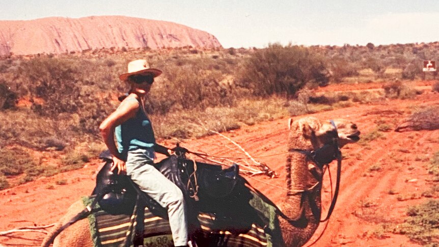 Jill Colwell, with wide-brimmed hat and jeans, sits on a camel surrounded by red dirt, and Uluru visible in the background.