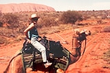 Jill Colwell, with wide-brimmed hat and jeans, sits on a camel surrounded by red dirt, and Uluru visible in the background.