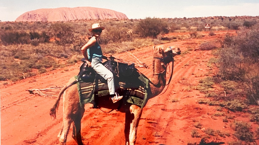 Jill Colwell, with wide-brimmed hat and jeans, sits on a camel surrounded by red dirt, and Uluru visible in the background.