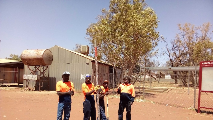 Four workers stand in a open field of red dust.  There are sheds in the background.