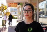Natalie Nguyen wears glasses and a black t-shirt while standing on the street in front of a lunch bar sign