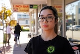 Natalie Nguyen wears glasses and a black t-shirt while standing on the street in front of a lunch bar sign