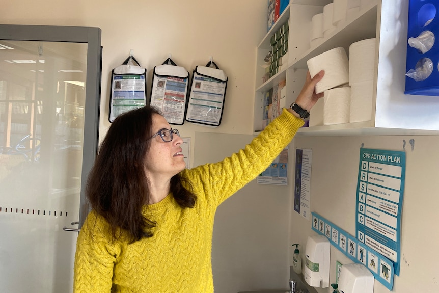 Woman reaches up to a shelf to get a roll of toilet paper.