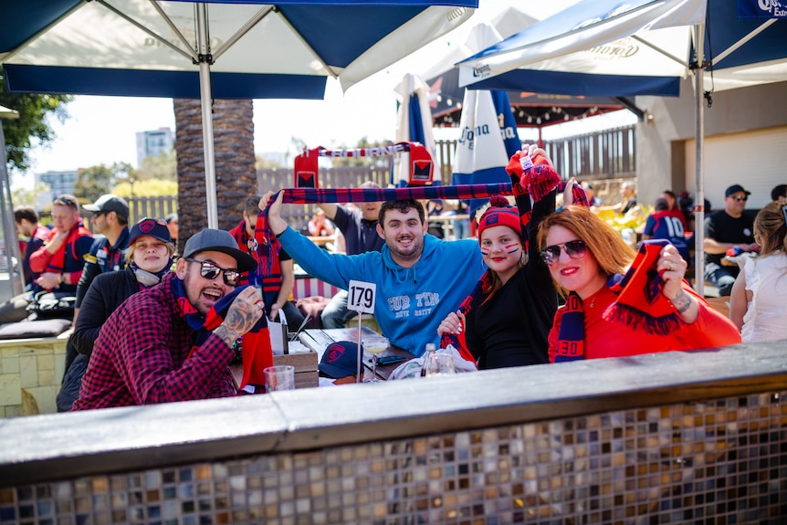 Five people sit at a pub table in a beer garden wearing Melbourne Demons merchandise.