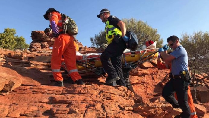 photo of SES volunteer in orange overalls and two police officers in uniform carrying unidentified person on stretcher over rock