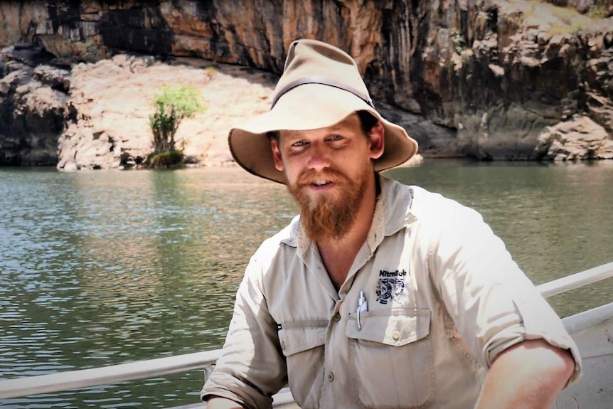 Bearded man wearing bush hat and tour guide wear posing for camera sitting in a metal boat in a river with dramatic rocks behind