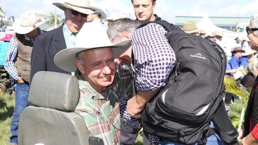 Man in a hat is hugged as he sits on a chair.