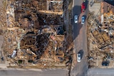 Drone image shows demolished buildings as cars drive down a road.