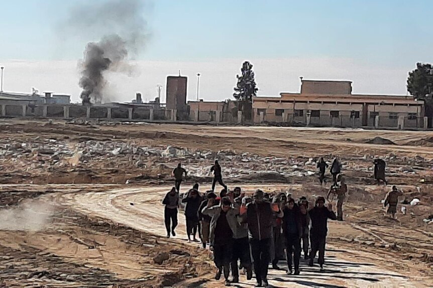 A group of men with their hands in the air walk across a barren area with a prison building in the background.