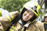A woman smiling, wearing a yellow firefighter's helmet and brown and yellow firefighter's jacket.