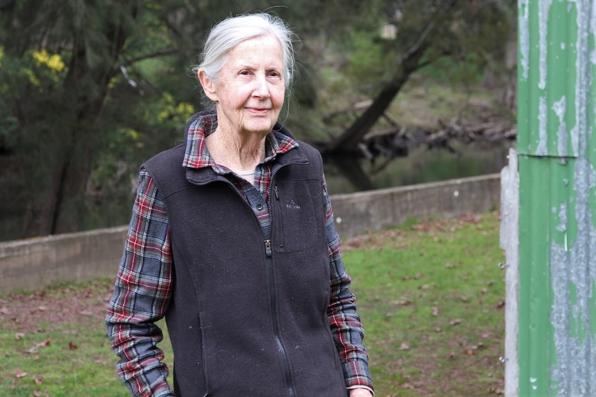 An elderly woman in a flannelette shirt and grey vest standing in front of a river. 