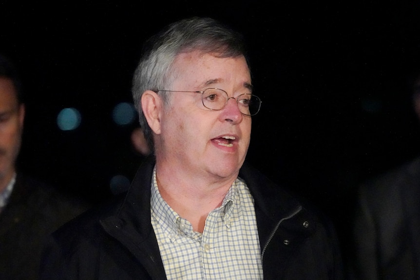 A frazzled-looking man with glasses in a checkered shirt speaks to reporters.